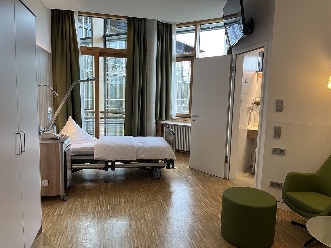 Ein Komfortpatientenzimmer mit Grünem Sessel, Bett, großen Fenstern und modernem Badezimmer.